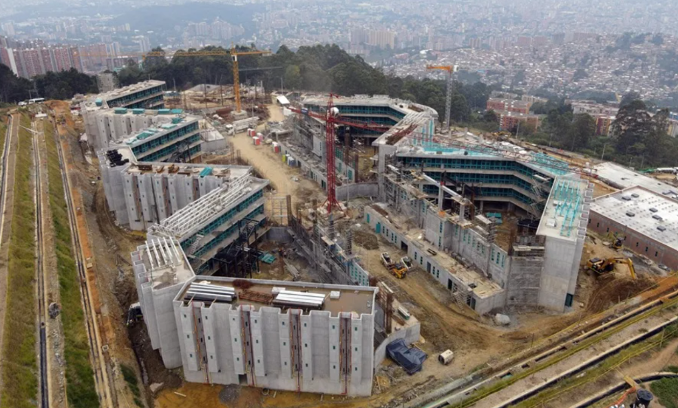 Aerial view of a large construction site with multiple connected concrete buildings, cranes, and construction equipment across a cleared site near a city edge.