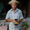 Man in a checked shirt and straw hat holds a phone showing a photo of a woman, amid rubble and debris in the background.