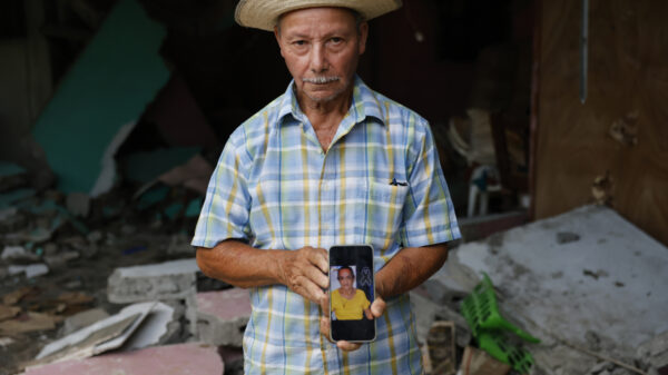 Man in a checked shirt and straw hat holds a phone showing a photo of a woman, amid rubble and debris in the background.