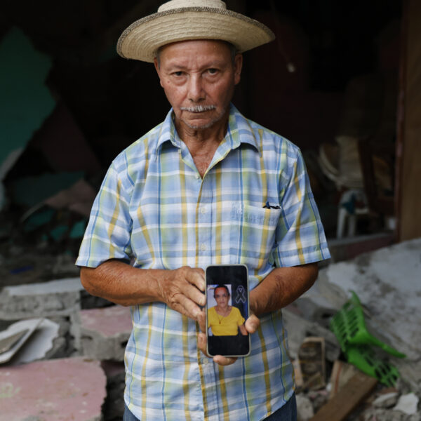 Man in a checked shirt and straw hat holds a phone showing a photo of a woman, amid rubble and debris in the background.