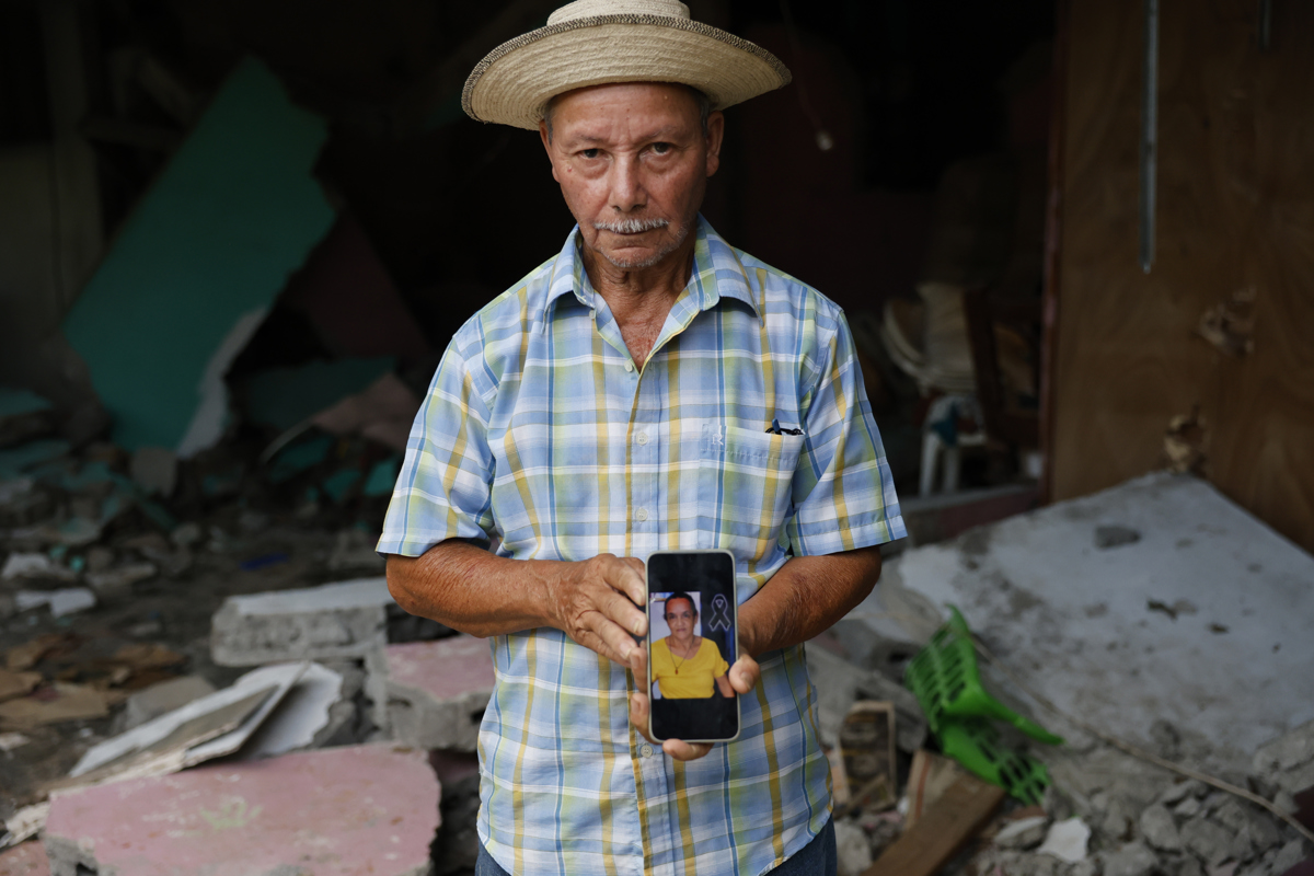 Man in a checked shirt and straw hat holds a phone showing a photo of a woman, amid rubble and debris in the background.