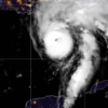 Infrared satellite image showing a tropical cyclone with a well-defined eye near the Gulf of Mexico, approaching the Florida coast with spiral rainbands over dark ocean waters and city lights along the coast visible at night.