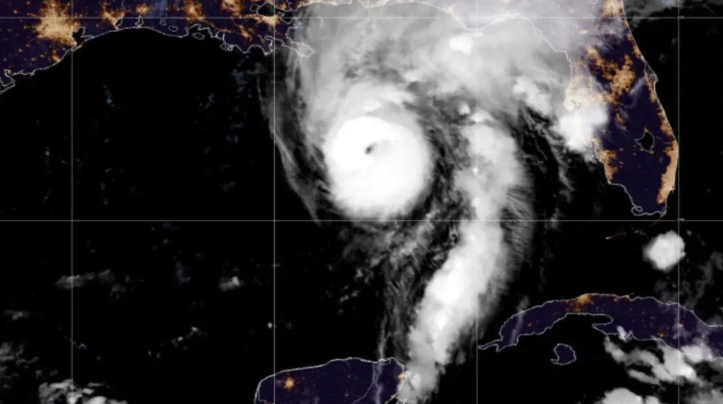 Infrared satellite image showing a tropical cyclone with a well-defined eye near the Gulf of Mexico, approaching the Florida coast with spiral rainbands over dark ocean waters and city lights along the coast visible at night.
