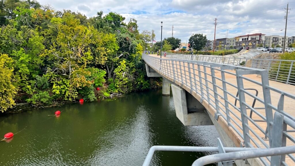 Pedestrian bridge over a green river with trees on the left and apartment buildings in the distance.