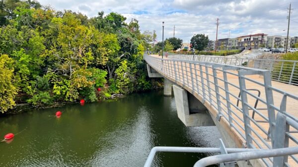 Pedestrian bridge over a green river with trees on the left and apartment buildings in the distance.