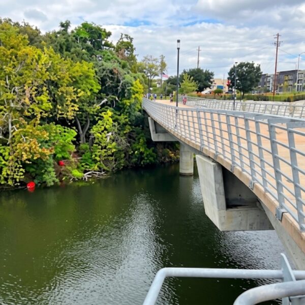 Pedestrian bridge over a green river with trees on the left and apartment buildings in the distance.