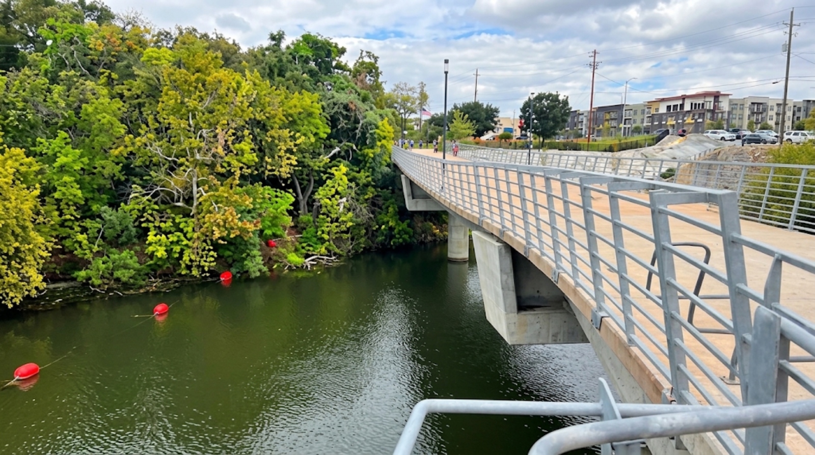 Pedestrian bridge over a green river with trees on the left and apartment buildings in the distance.