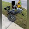 Motorcycle crash scene: a black bike on the grassy verge beside a sidewalk, with responders in high‑visibility gear tending to a rider; helmet sits nearby.