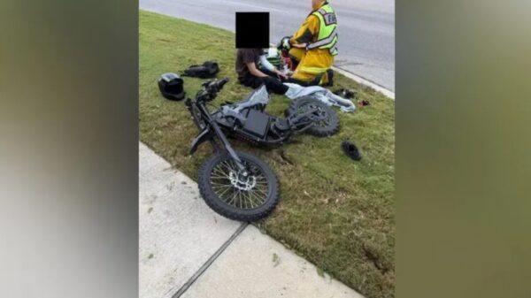 Motorcycle crash scene: a black bike on the grassy verge beside a sidewalk, with responders in high‑visibility gear tending to a rider; helmet sits nearby.