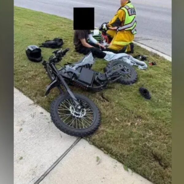 Motorcycle crash scene: a black bike on the grassy verge beside a sidewalk, with responders in high‑visibility gear tending to a rider; helmet sits nearby.