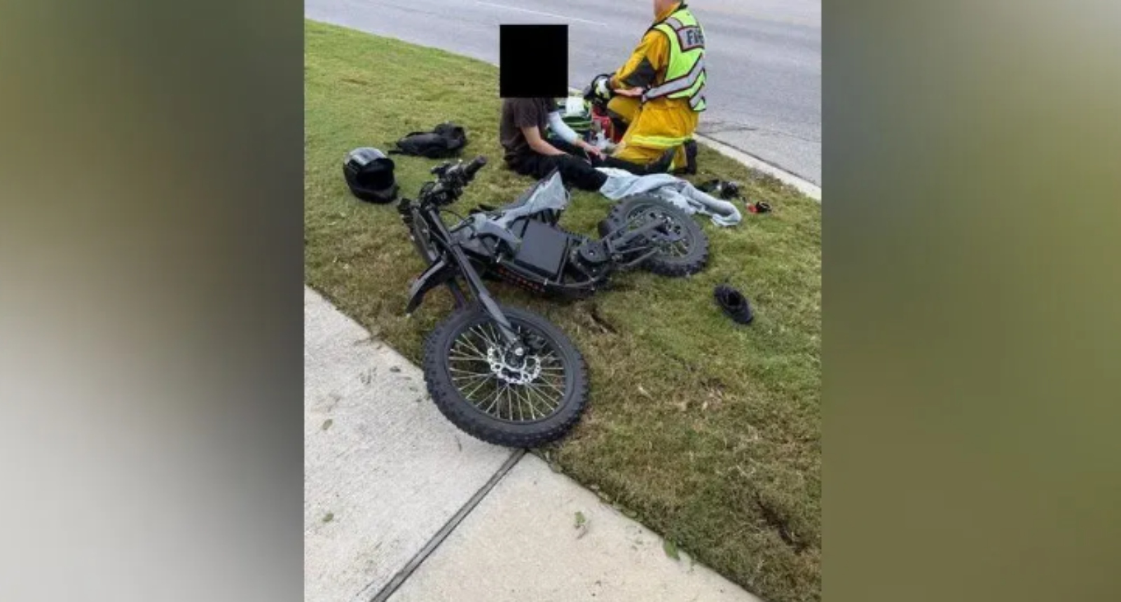 Motorcycle crash scene: a black bike on the grassy verge beside a sidewalk, with responders in high‑visibility gear tending to a rider; helmet sits nearby.
