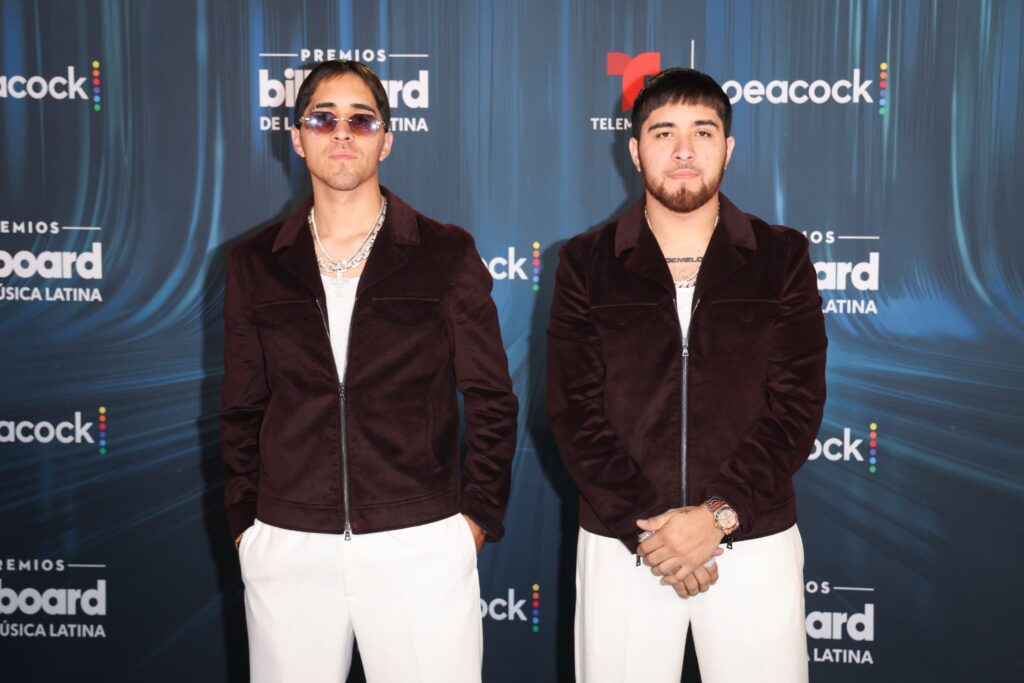 Two men pose in matching maroon jackets and white pants at a Billboard Latin music awards backdrop with logos behind them.