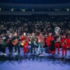 Band and brass section perform on stage in front of a cheering crowd holding up illuminated phones at night concert venue.