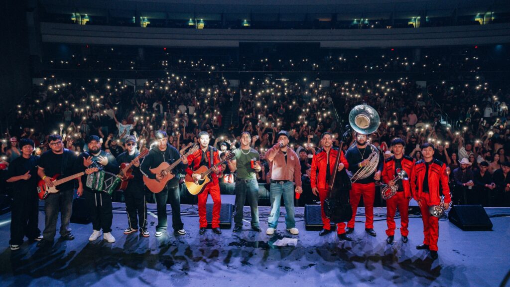 Band and brass section perform on stage in front of a cheering crowd holding up illuminated phones at night concert venue.