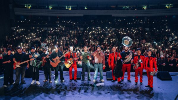 Band and brass section perform on stage in front of a cheering crowd holding up illuminated phones at night concert venue.