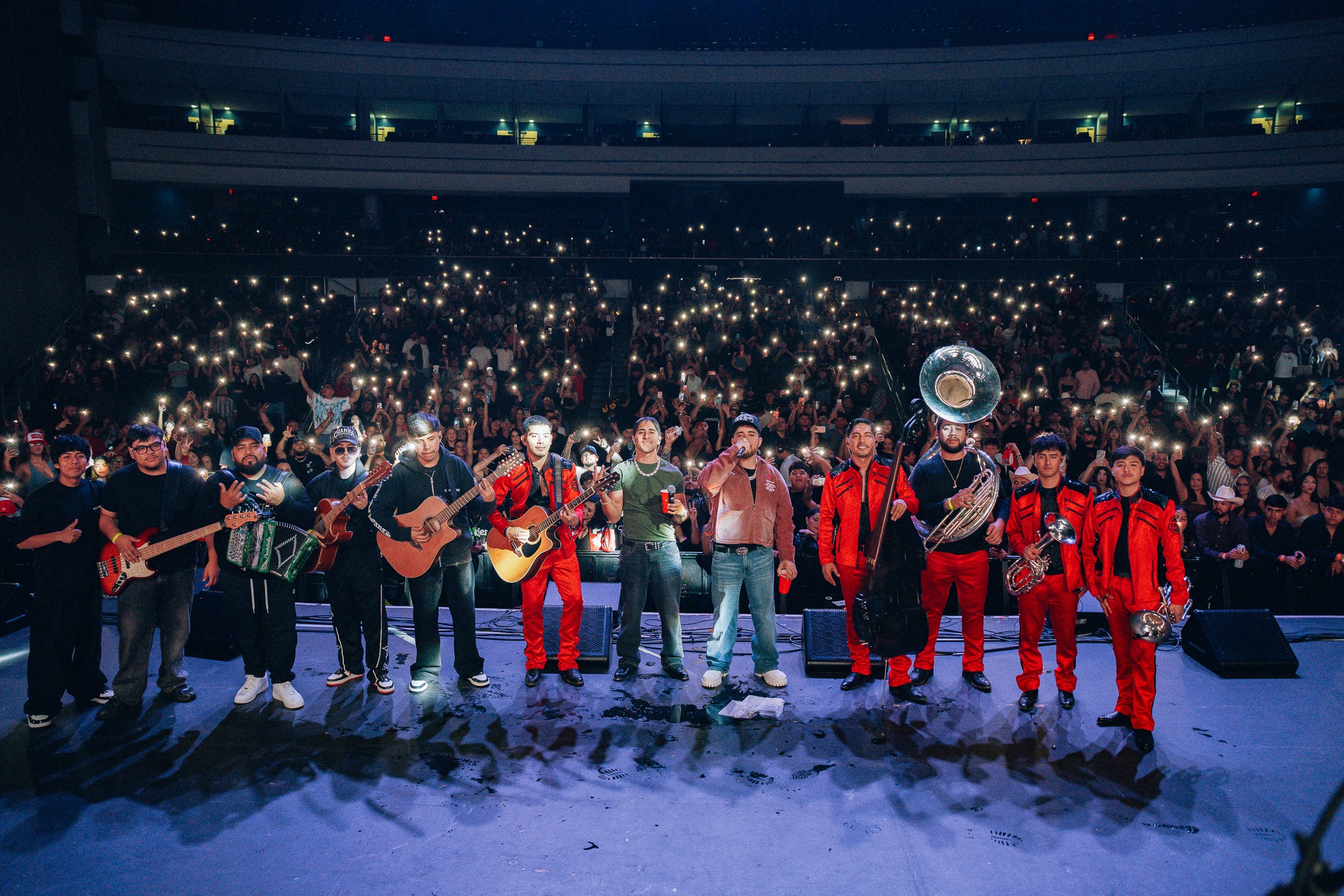 Band and brass section perform on stage in front of a cheering crowd holding up illuminated phones at night concert venue.