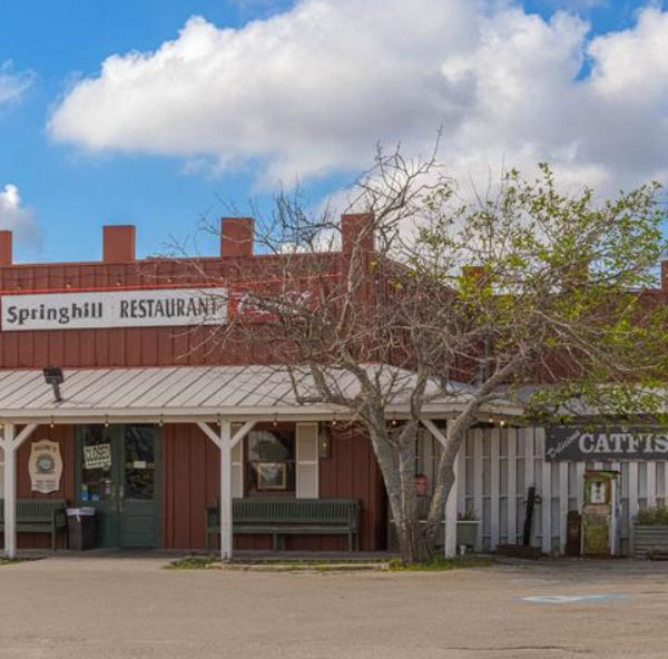 Red wooden restaurant with a covered porch and signs reading Springhill Restaurant, Coca-Cola, and Catfish & Shrimp menu under a blue sky.