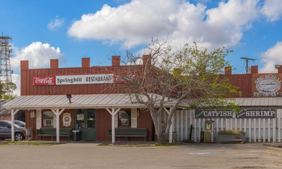 Red wooden restaurant with a covered porch and signs reading Springhill Restaurant, Coca-Cola, and Catfish & Shrimp menu under a blue sky.