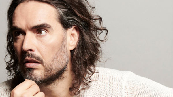 Close-up of a man with long wavy hair and stubble, resting his chin and looking to the left.