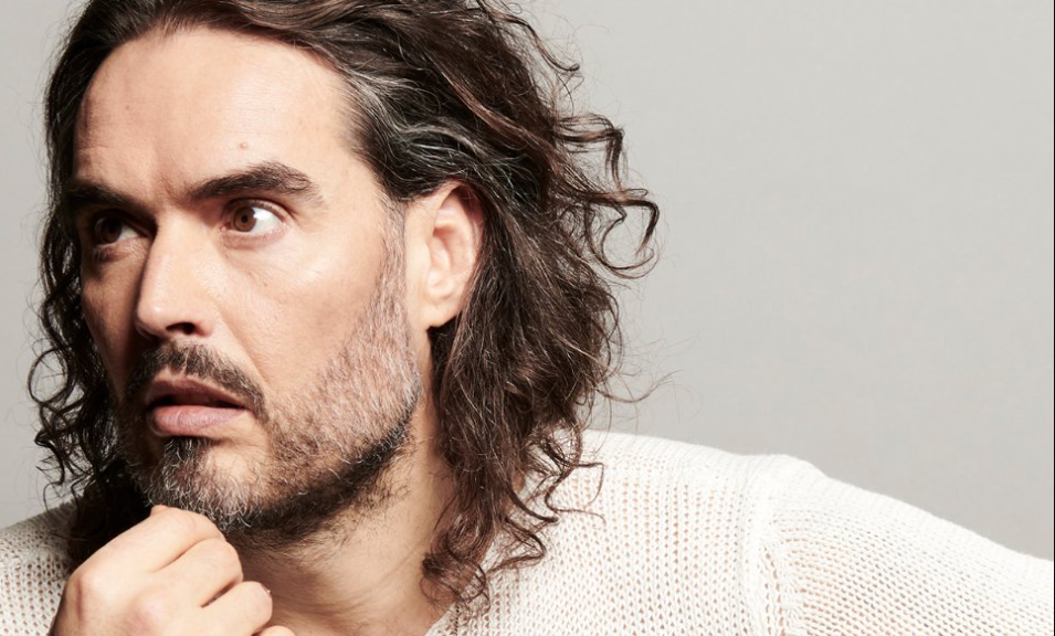 Close-up of a man with long wavy hair and stubble, resting his chin and looking to the left.