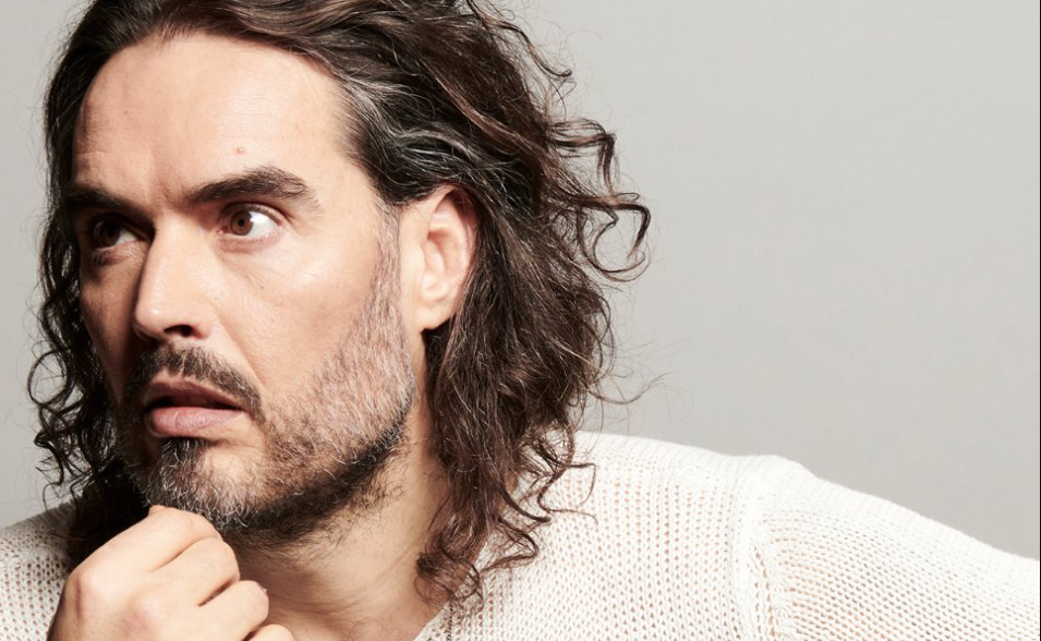Close-up of a man with long wavy hair and stubble, resting his chin and looking to the left.