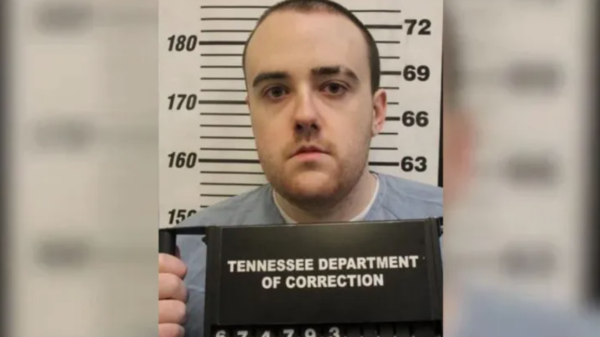Mugshot portrait of a man in a gray shirt in front of a height measurement board, holding a Tennessee Department of Correction sign below.