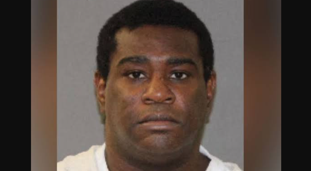 Mugshot of a Black man with short black hair wearing a white shirt against a gray backdrop with a neutral expression.