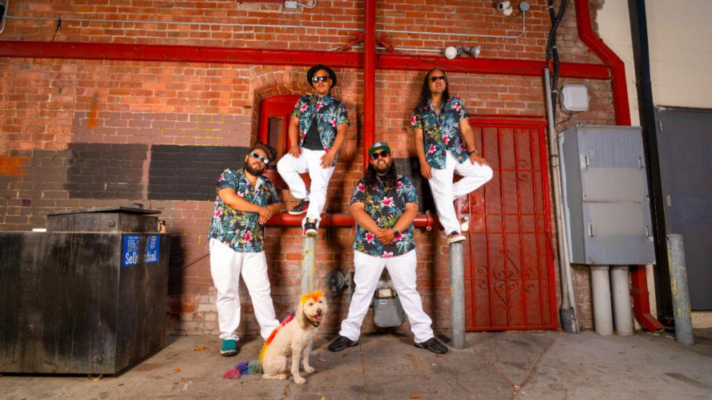 Five men in tropical shirts and white pants pose against a brick wall with red pipes; a small rainbow-colored dog sits at their feet.