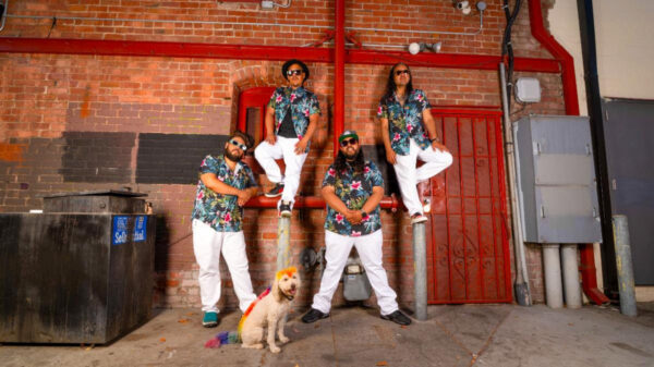 Five men in tropical shirts and white pants pose against a brick wall with red pipes; a small rainbow-colored dog sits at their feet.