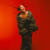 Joyful portrait of a person in a bulky dark green puffer jacket, laughing and dancing against a bright orange background.