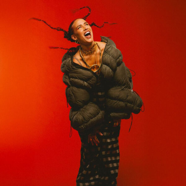 Joyful portrait of a person in a bulky dark green puffer jacket, laughing and dancing against a bright orange background.