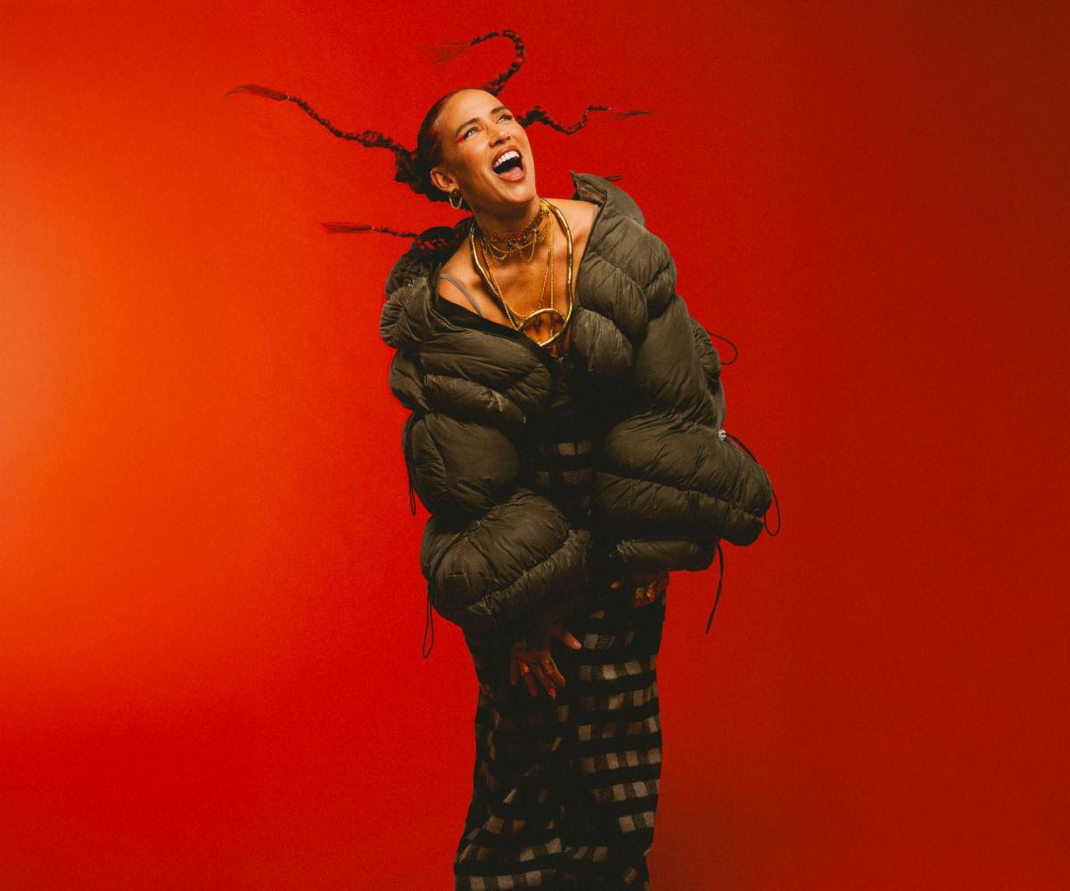Joyful portrait of a person in a bulky dark green puffer jacket, laughing and dancing against a bright orange background.