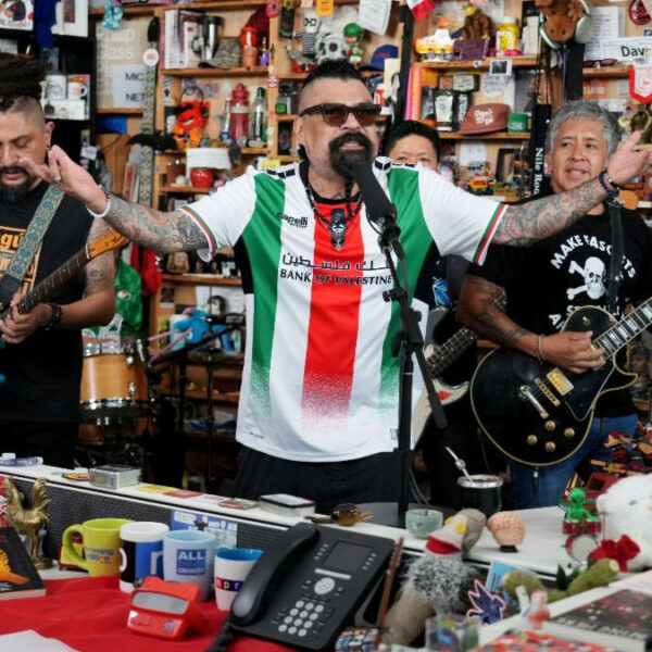 Man with arms outstretched singing into a microphone while a small band plays in a busy toy-and-gift shop behind him
