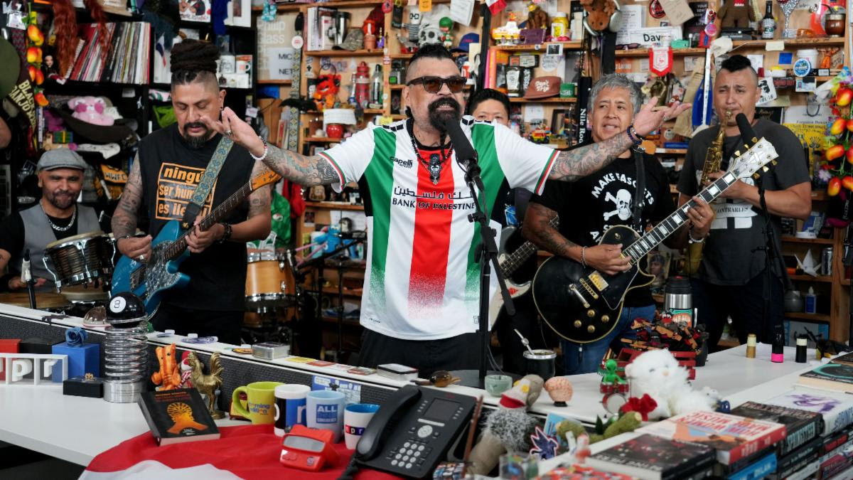Man with arms outstretched singing into a microphone while a small band plays in a busy toy-and-gift shop behind him