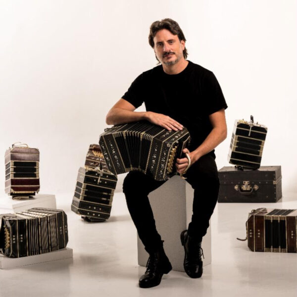 Male musician in a black shirt sits on a white block, playing a large accordion, surrounded by several other accordions in a studio.