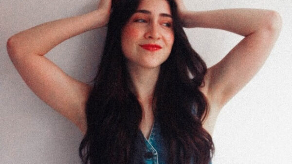 Young woman with long dark hair wearing a sleeveless denim top and jeans, arms raised behind her head, smiling against a light wall.