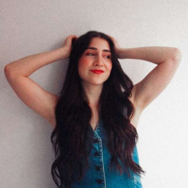 Young woman with long dark hair wearing a sleeveless denim top and jeans, arms raised behind her head, smiling against a light wall.
