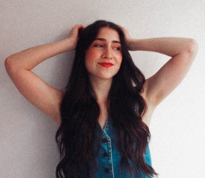 Young woman with long dark hair wearing a sleeveless denim top and jeans, arms raised behind her head, smiling against a light wall.