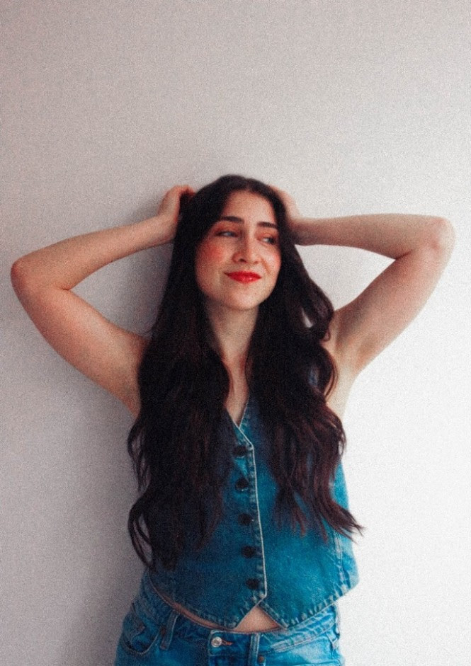 Young woman with long dark hair wearing a sleeveless denim top and jeans, arms raised behind her head, smiling against a light wall.