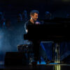 Pianist smiling while playing a grand piano on a dimly lit stage with an orchestra in the background and blue stage lighting, creating a smoky atmosphere.