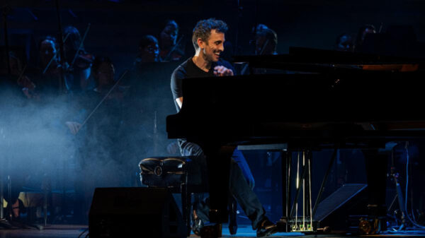 Pianist smiling while playing a grand piano on a dimly lit stage with an orchestra in the background and blue stage lighting, creating a smoky atmosphere.