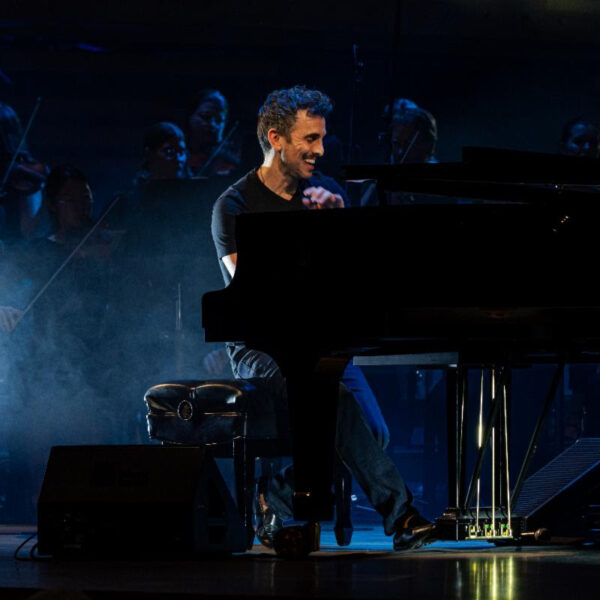 Pianist smiling while playing a grand piano on a dimly lit stage with an orchestra in the background and blue stage lighting, creating a smoky atmosphere.