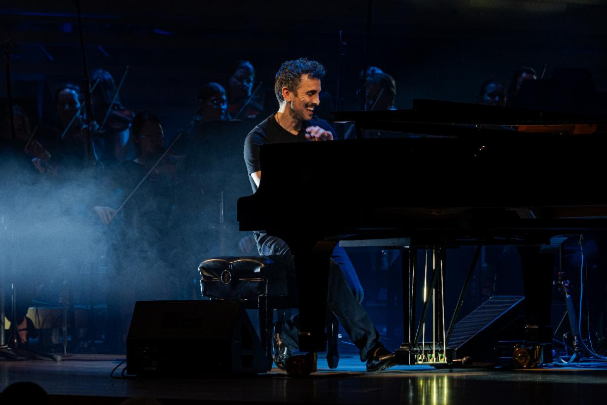 Pianist smiling while playing a grand piano on a dimly lit stage with an orchestra in the background and blue stage lighting, creating a smoky atmosphere.